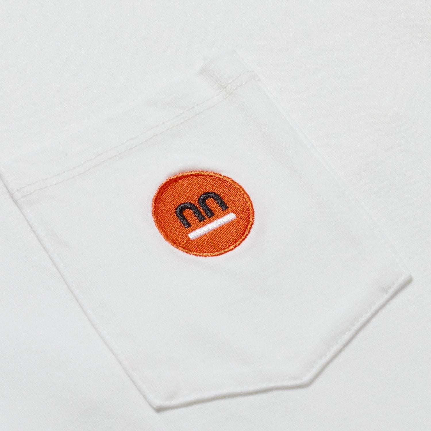 Close-up of the white t-shirt's pocket highlighting the orange Pamuuc logo on an embroidered patch.